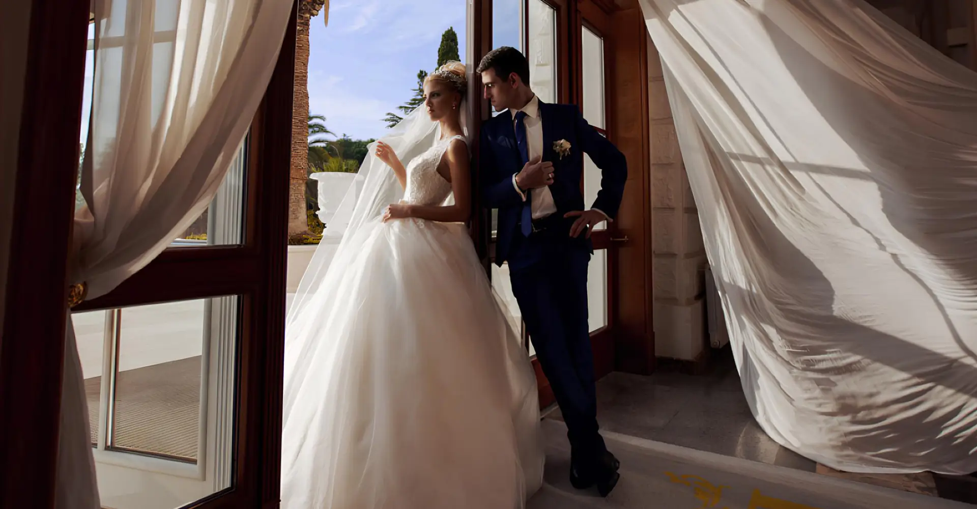 The bride in a fluffy white dress and the groom pose at the open door of the balcony The bride and groom pose on the balcony