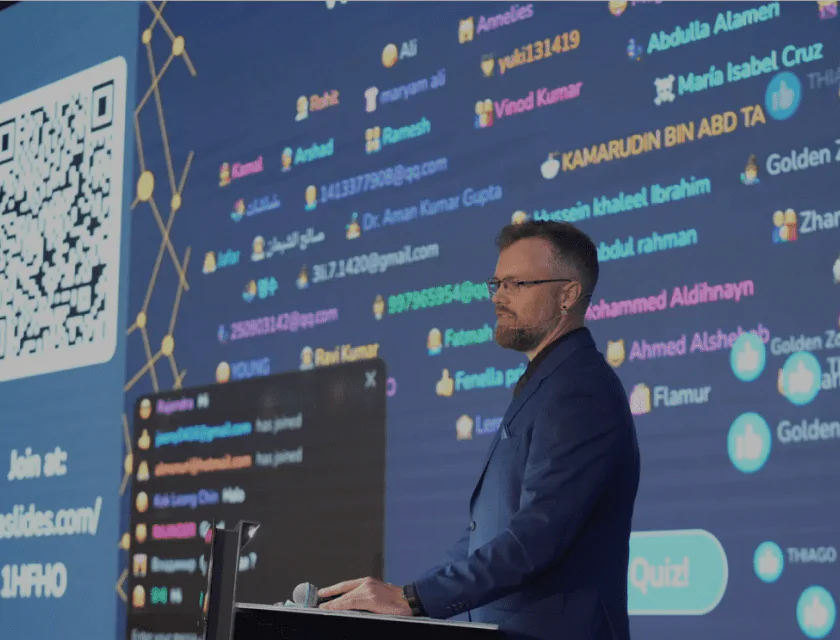 A man with glasses and a microphone is speaking at a conference, with a screen with the names of the participants in the background A man with glasses speaks at a conference
