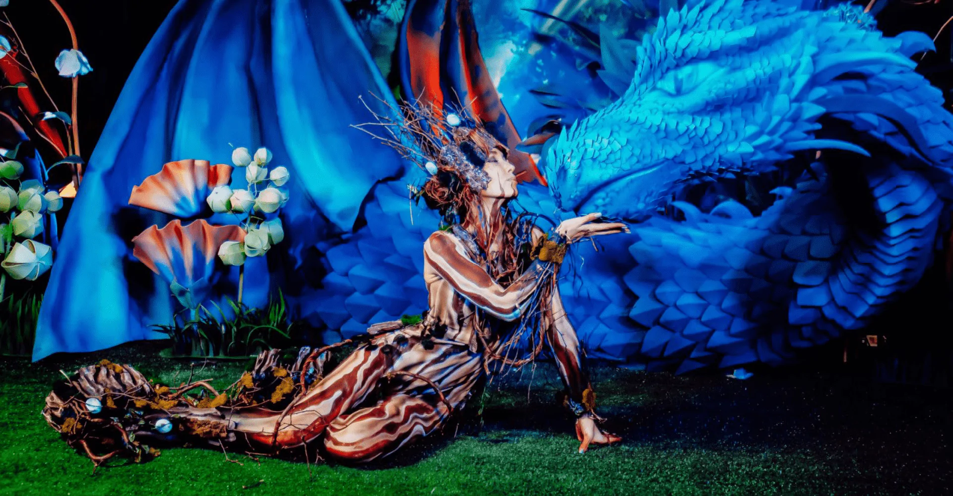 A girl dressed as a fabulous creature is going to kiss a sculpture of a blue dragon A fabulous creature kisses a blue dragon