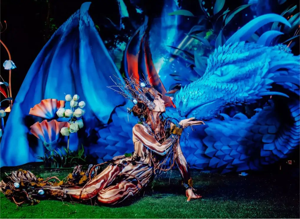 A girl dressed as a fairy-tale character is going to kiss a sculpture of a blue dragon A fabulous creature kisses a blue dragon