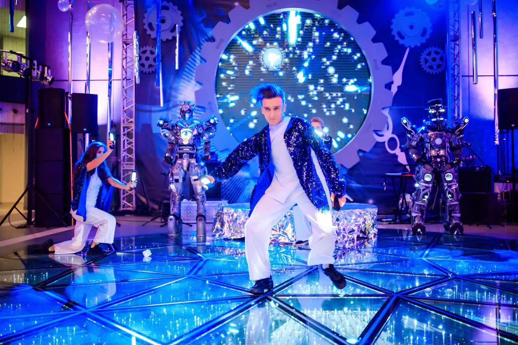 A dancer performs a number on a glass floor, with two people in robot costumes standing in the background A man is dancing on a glass floor