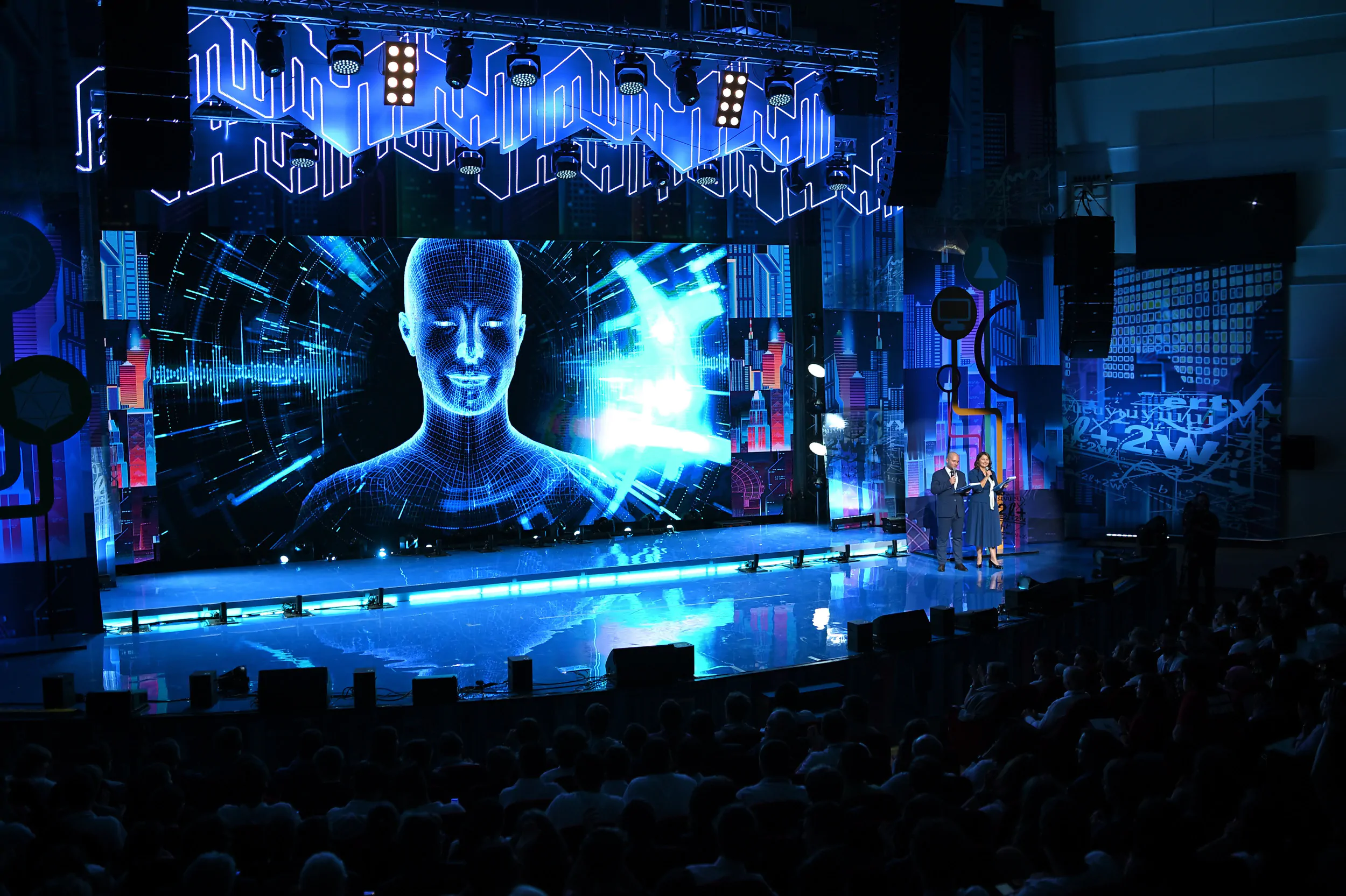 Two hosts on stage with futuristic image on screen